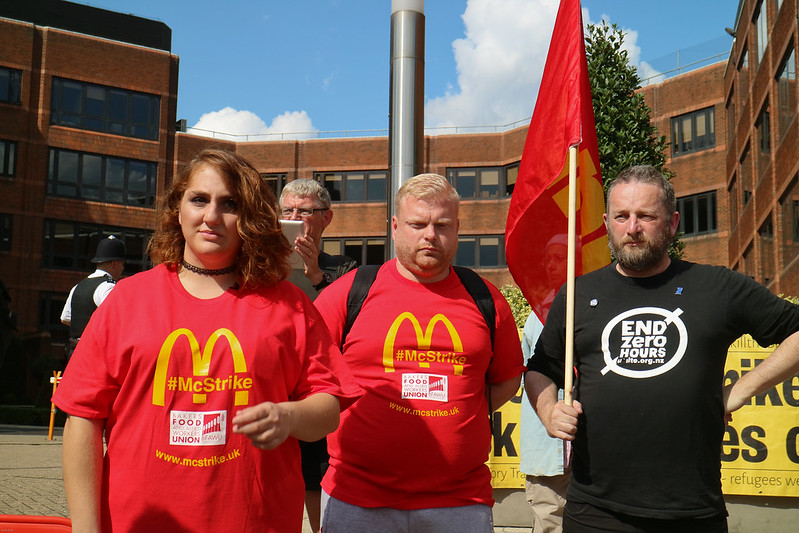 Image shows three striking McDonald's workers, on a sunny day, one holding a red flag and wearing a T-shirt that reads 'End Zero Hours Contracts'