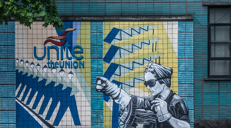 Image shows a mural on the side of Unite's Transport House of a woman at a loom and a row of stylised workers. The Unite the Union logo is included. The picture is painted in blues and yellows.