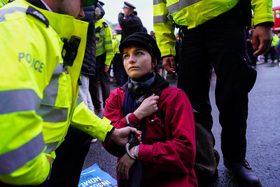 Image shows a woman wearing red kneeling on the ground in handcuffs, being pulled to her feet by a police officer, from a climate protest in 2022.