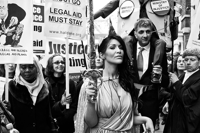 Image shows a woman dressed as Lady Justice in a protest amongst placards that read 